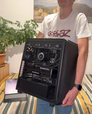 A person holding an open black desktop PC with visible cooling and GeForce RTX graphics card, standing in a cozy room with a striped bed cover, green plant, and a laptop in the background.