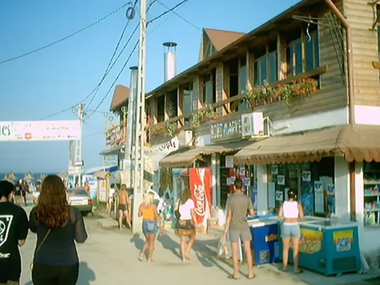 People walking down Vama Veche’s main street near the beach in 2003, passing wooden houses, shops, and bars.