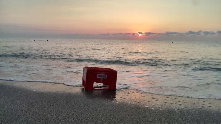 Red Ursus beer crate washed onto the shore during sunrise, waves gently touching it.