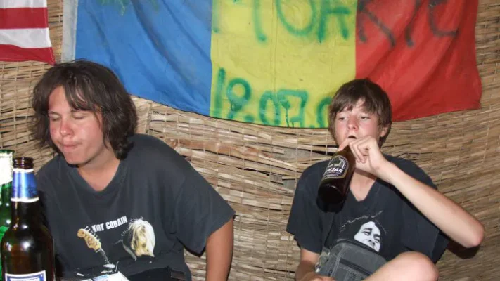 Two young men sit inside a rustic shack, drinking beer beneath a Romanian flag tagged with graffiti.