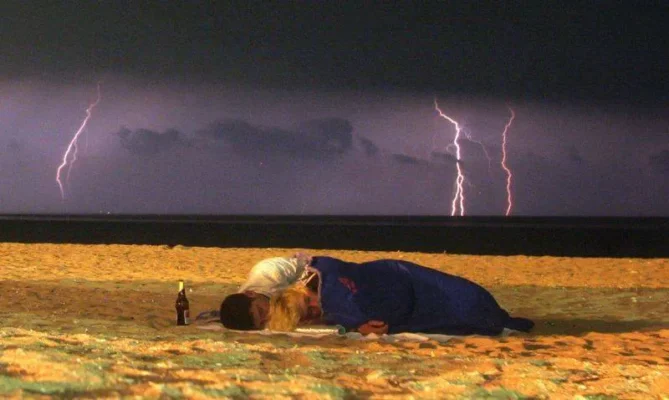 Two people sleeping on the beach under a blanket with lightning in the distance.