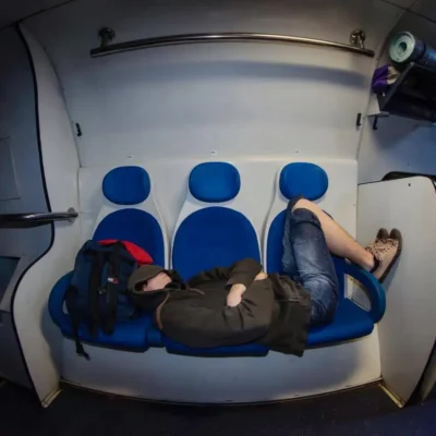 A young traveler sleeping stretched across three blue train seats at night, hoodie over the face, backpack as a pillow, on the cheap 2 AM Bucharest–Mangalia train.