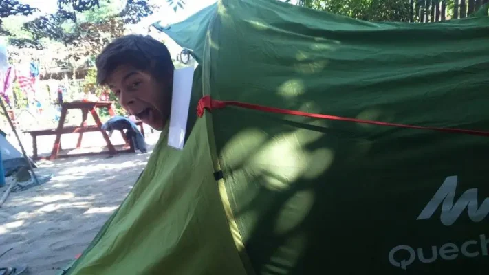 A young man poking his head out of a green Quechua tent with a wide, playful expression, at a campsite with sand and picnic tables.