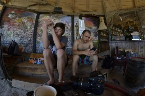 Two young men resting inside a beach bar in Vama Veche, one looking tired, the other on his phone, with colorful murals behind them.