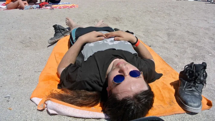 A young man with long hair and blue round sunglasses lies on an orange towel on the beach in Vama Veche, boots placed beside him in the sand.