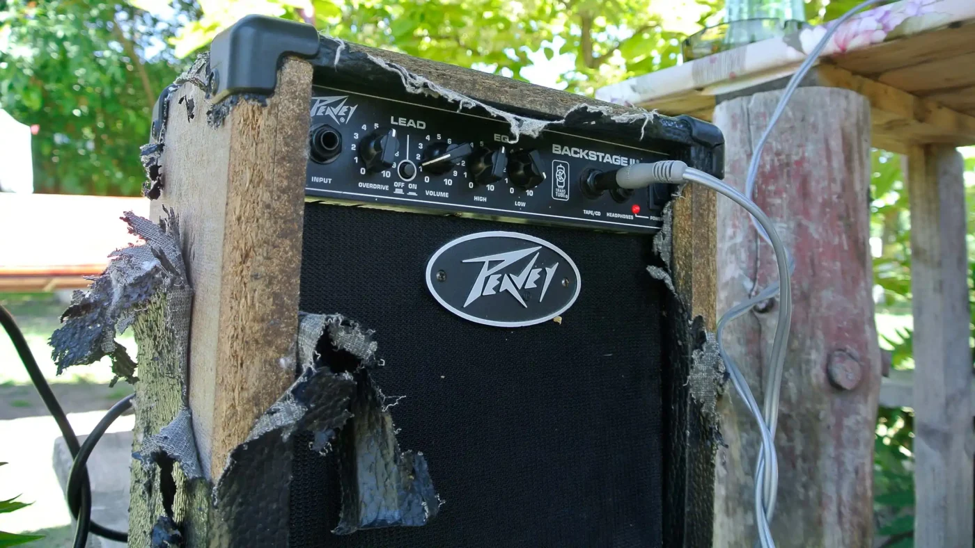 A weathered Peavey Backstage guitar amplifier with torn fabric and peeling edges, patched into a makeshift outdoor setup.