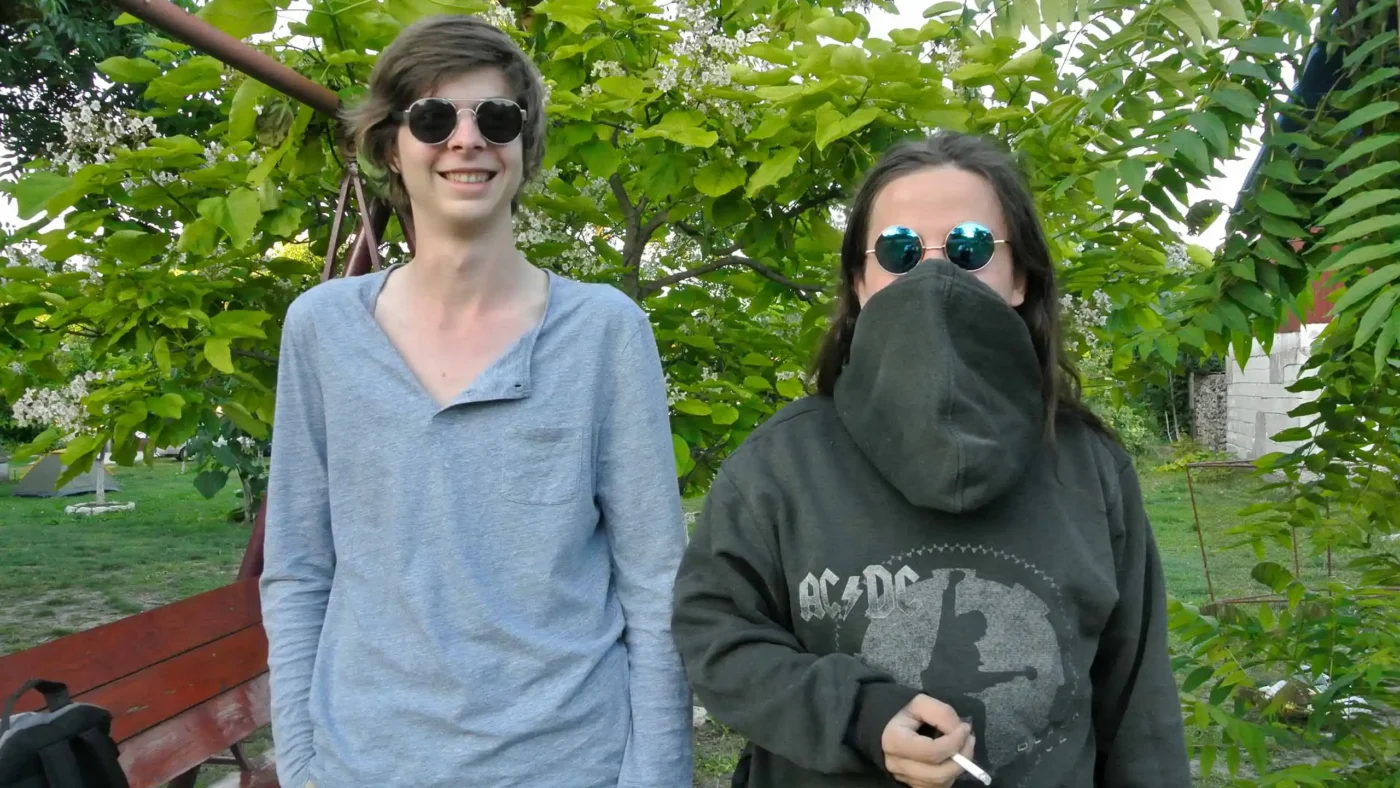 Two friends posing in the campsite, one grinning wide, the other with a hoodie pulled over their face, cigarette in hand, both wearing round sunglasses.