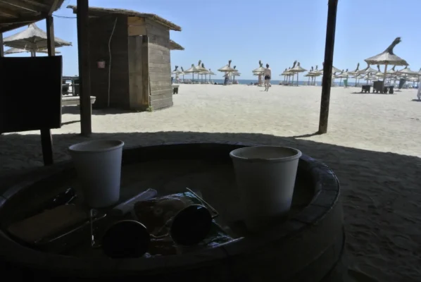 Two plastic cups on a barrel table inside a shaded beach bar, looking out toward straw umbrellas and the sea in Vama Veche.
