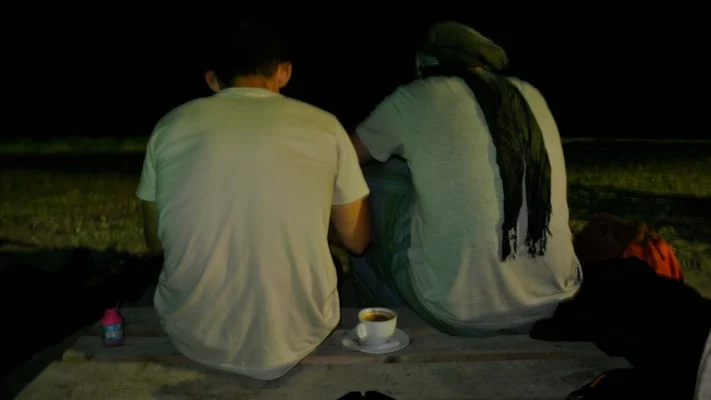 Two people sitting side by side on a wooden bench at night, one with a scarf, a cup of coffee between them.