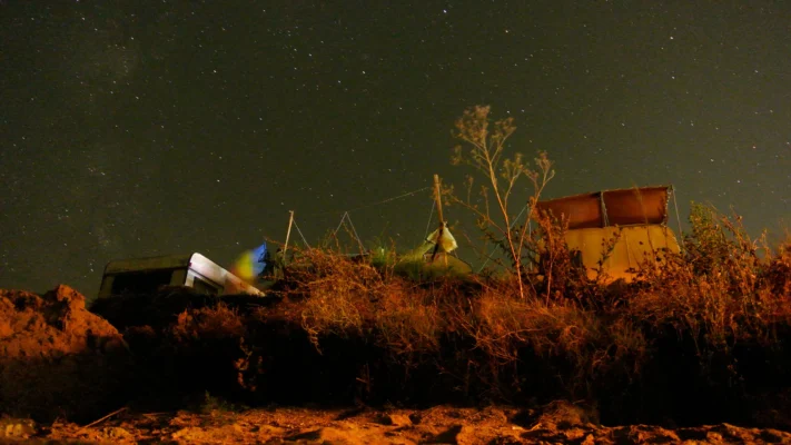 Long-exposure view from the beach of tents and a shack silhouetted on the cliff edge beneath a star-filled sky in Vama Veche.
