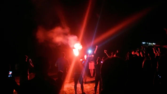 Crowd at night on the beach, one person holding a bright red flare with smoke rising.