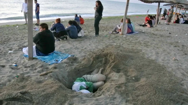 Person asleep in a dug-out hole on the beach at dawn, surrounded by people sitting on blankets.