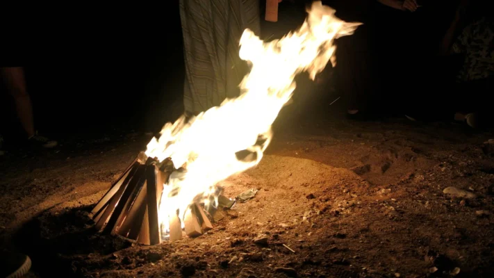 Campfire burning brightly on the beach at night with people standing around.