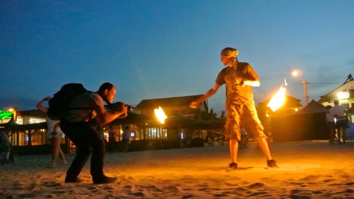Fire performer on the beach at dusk, holding flaming poi as a cameraman films closely.