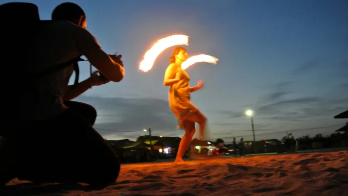 Female fire dancer twirling poi on the sand, flames glowing against the twilight sky.