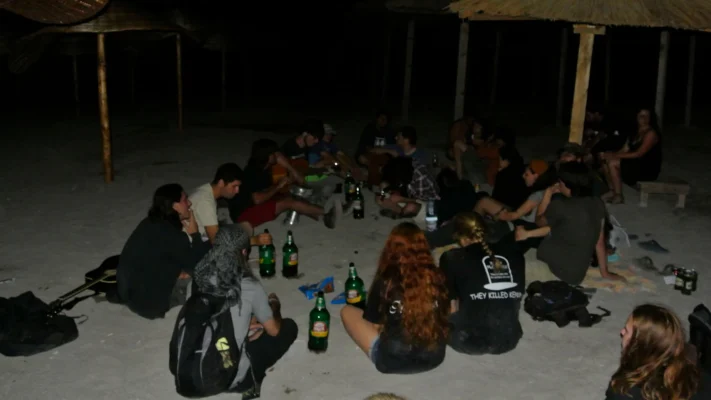Group of friends sitting on the beach at night with guitars and drinks under straw umbrellas.