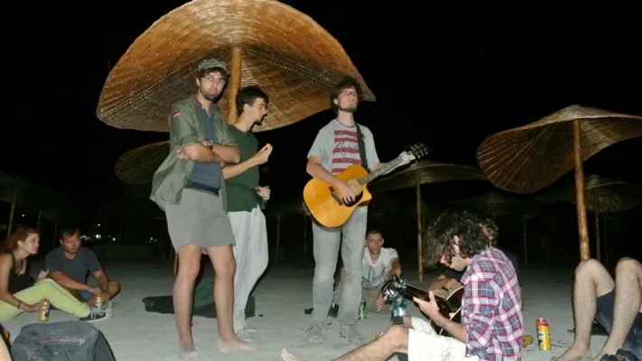 Man standing with a guitar among friends under straw umbrellas, playing music on the beach at night.