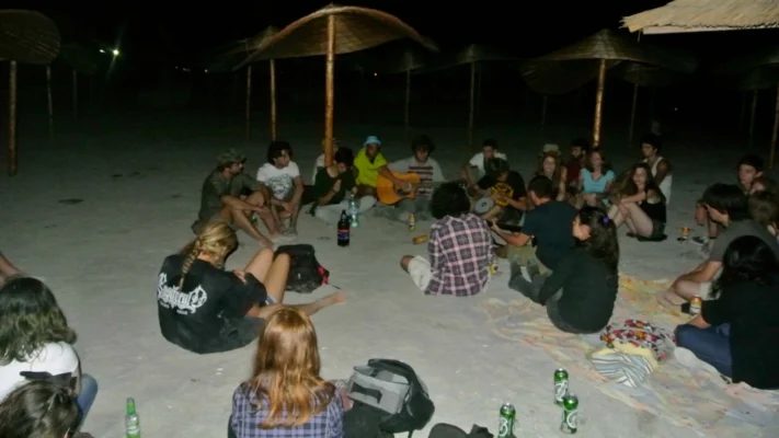 Large group of young people sitting in a circle on the sand at night, playing guitars and drinking.