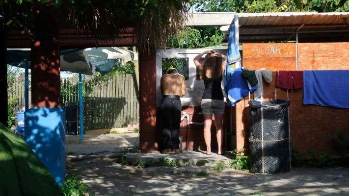 A man and a woman standing shirtless at the entrance of a Vama camping restroom marked "PISOAR," a shaded spot in the summer light.