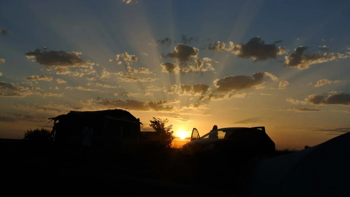Golden sunset with sun rays breaking through scattered clouds, silhouetting a small shack and a parked car.