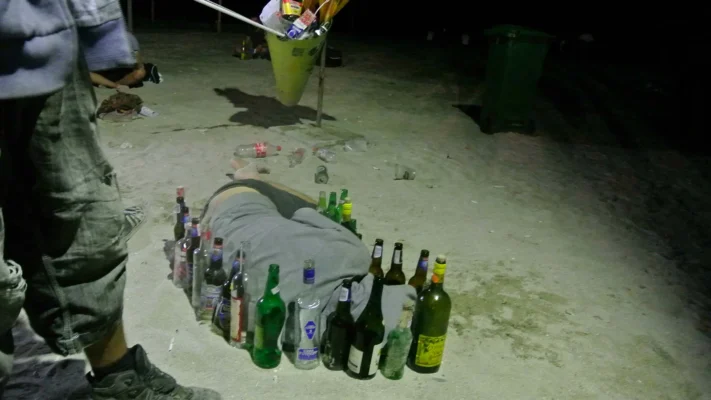 Man lying face down on the sand, surrounded by a wall of empty glass bottles.