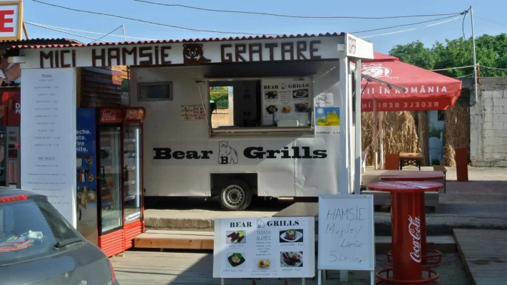 A food stall in Vama Veche named "Bear Grills," offering mici, hamsie, and grilled food, with handwritten signs advertising cheap meals like fried anchovies for 5 lei.