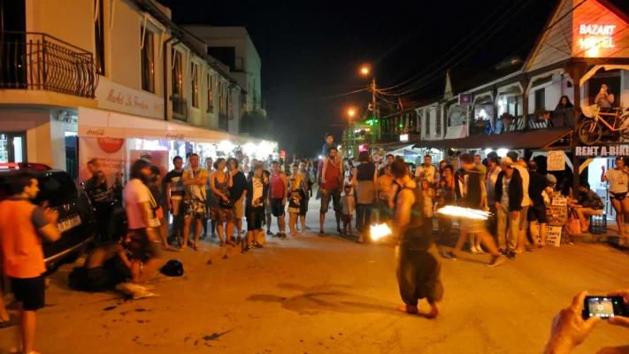 Fire dancer spinning flaming poi in the street at night, car headlights and a crowd in the background.