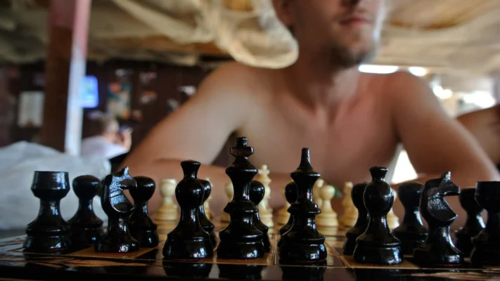 Close-up of black chess pieces on a board, a shirtless player blurred in the background.