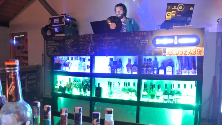 DJ standing behind a small lit-up bar, chalkboard drinks menu above him.