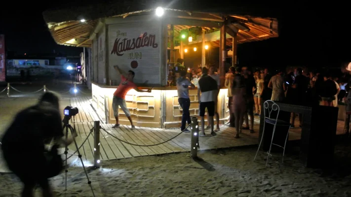 Wooden beach bar at night, lit warmly, with people gathered around.