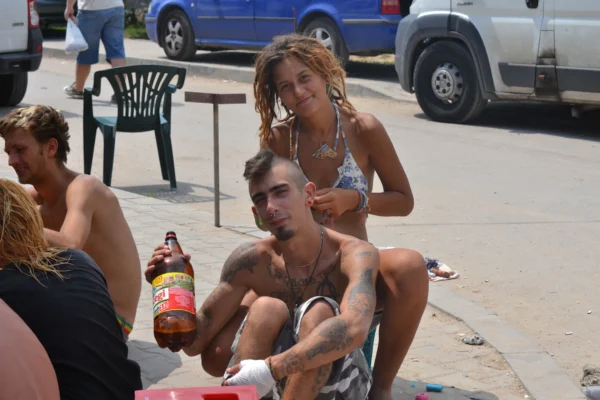 A young couple in Vama Veche, with tattoos, piercings, and dreadlocks, sitting in the sun with a large beer.