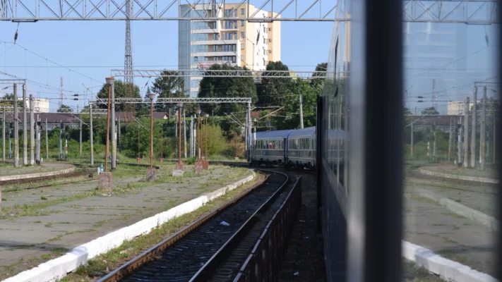 A Romanian passenger train curving out of a station, with overhead wires, empty platforms, and a tall apartment block in the background.