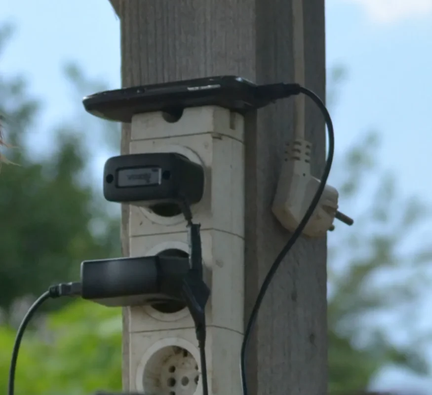 A cluster of chargers plugged into an old outdoor power strip nailed to a wooden post, phones and devices charging in the open air.