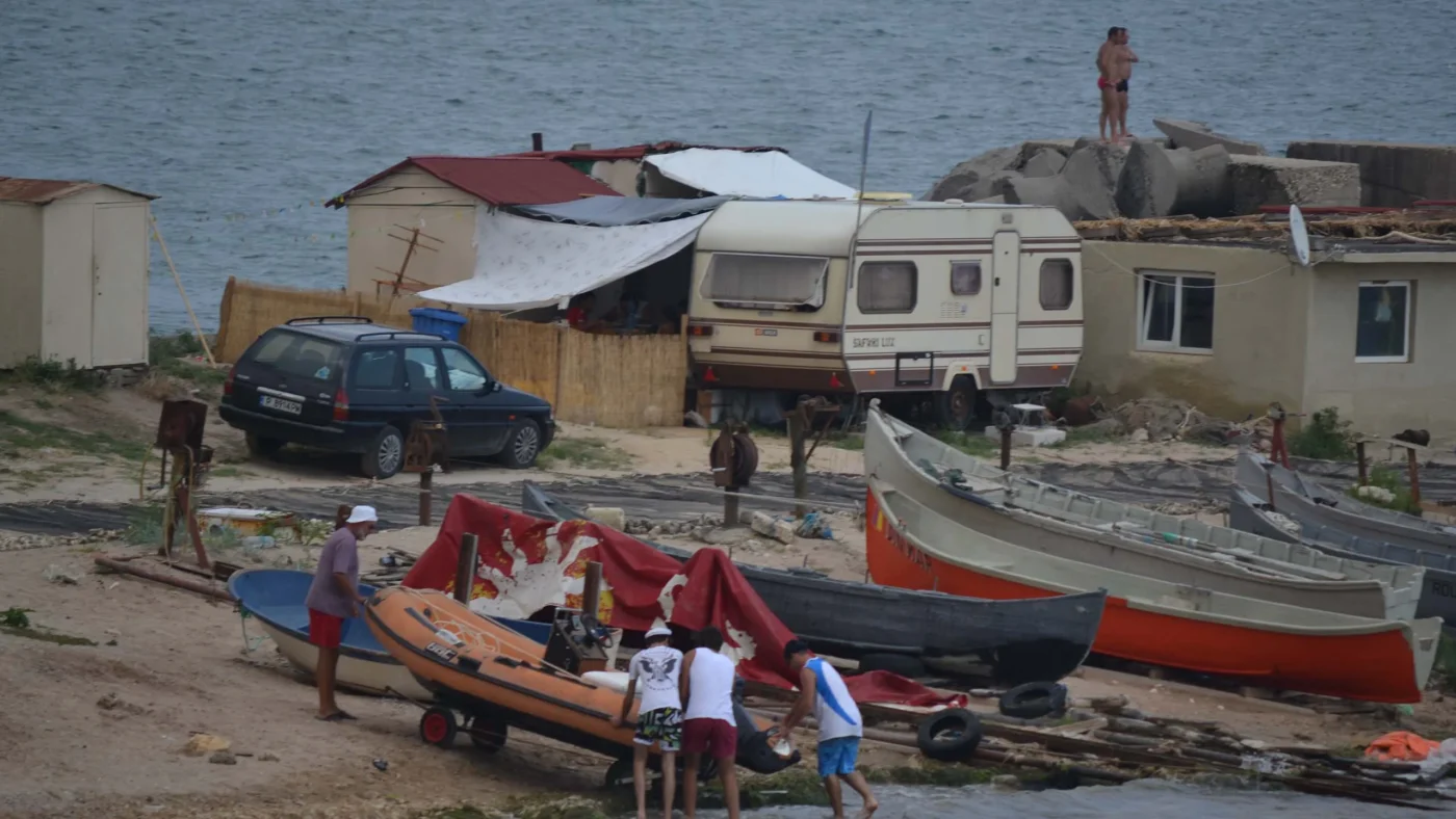 Fishermen's homes in 2 Mai, Romania, with small boats on the shore, a caravan, and locals working by the water.