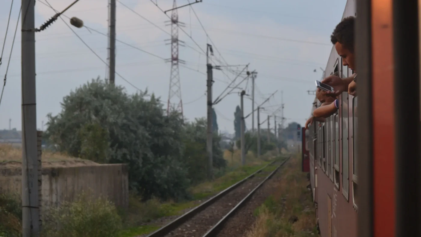 People leaning out of the windows of a Romanian train, checking phones and watching the tracks as the train passes through power lines and countryside.