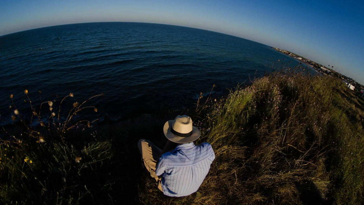 Person in a light hat sitting on the cliff, looking out over a round horizon of blue sea.