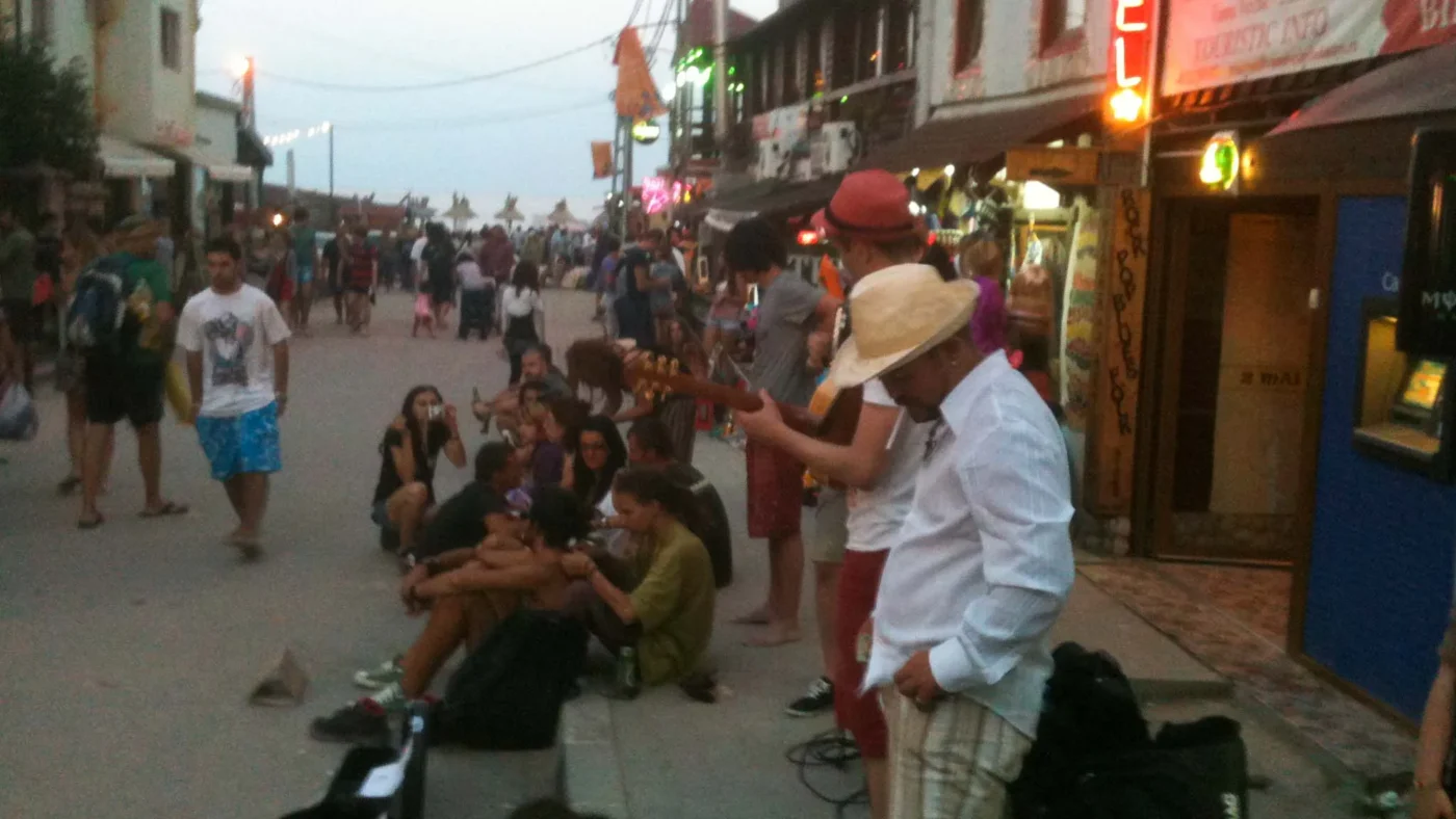 Street scene in Vama Veche at dusk - a crowd gathers on the curb, bottles in hand, listening to two guitarists playing under the neon lights of the main strip.