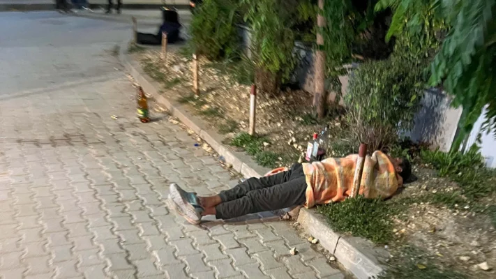 A person lies on the pavement wrapped in a blanket beside empty alcohol bottles.