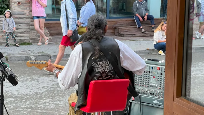 Jaguarul A long-haired man in a leather vest plays electric guitar on a busy street with an audio mixer.
