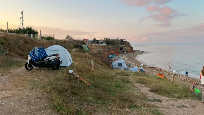 A scooter and tents stand on a grassy cliff above the beach at sunset.