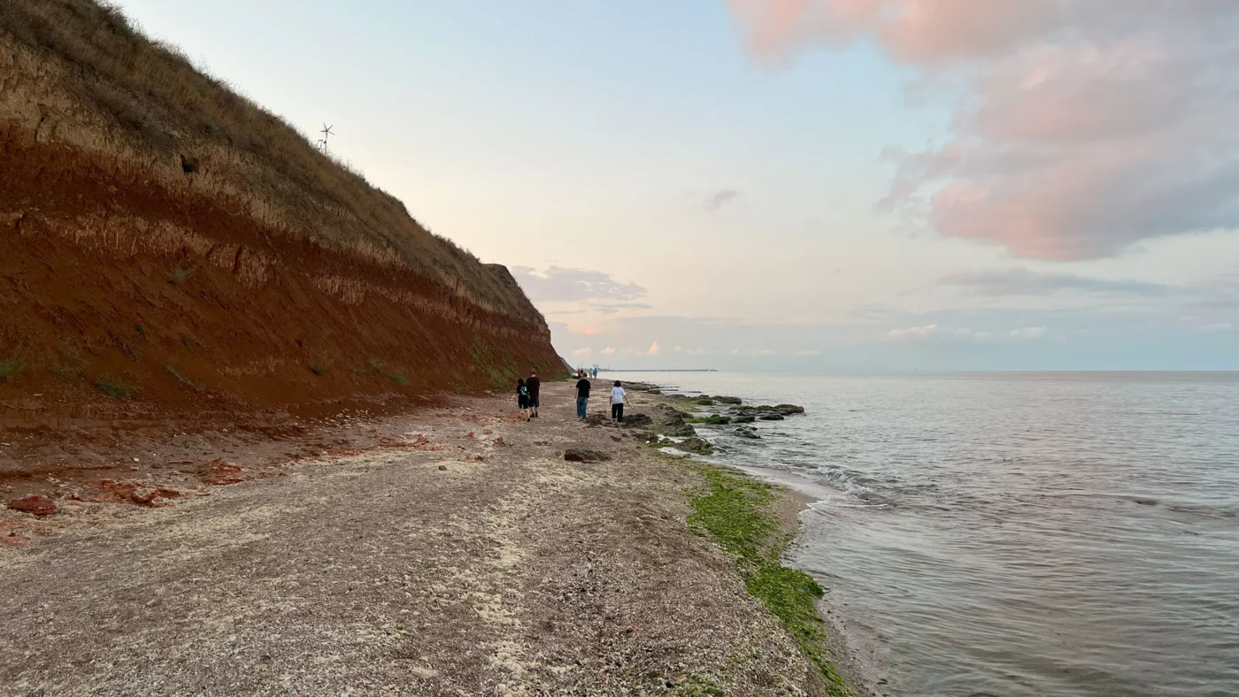 People walking along the rocky shoreline beneath steep red cliffs at sunset in Vama Veche.