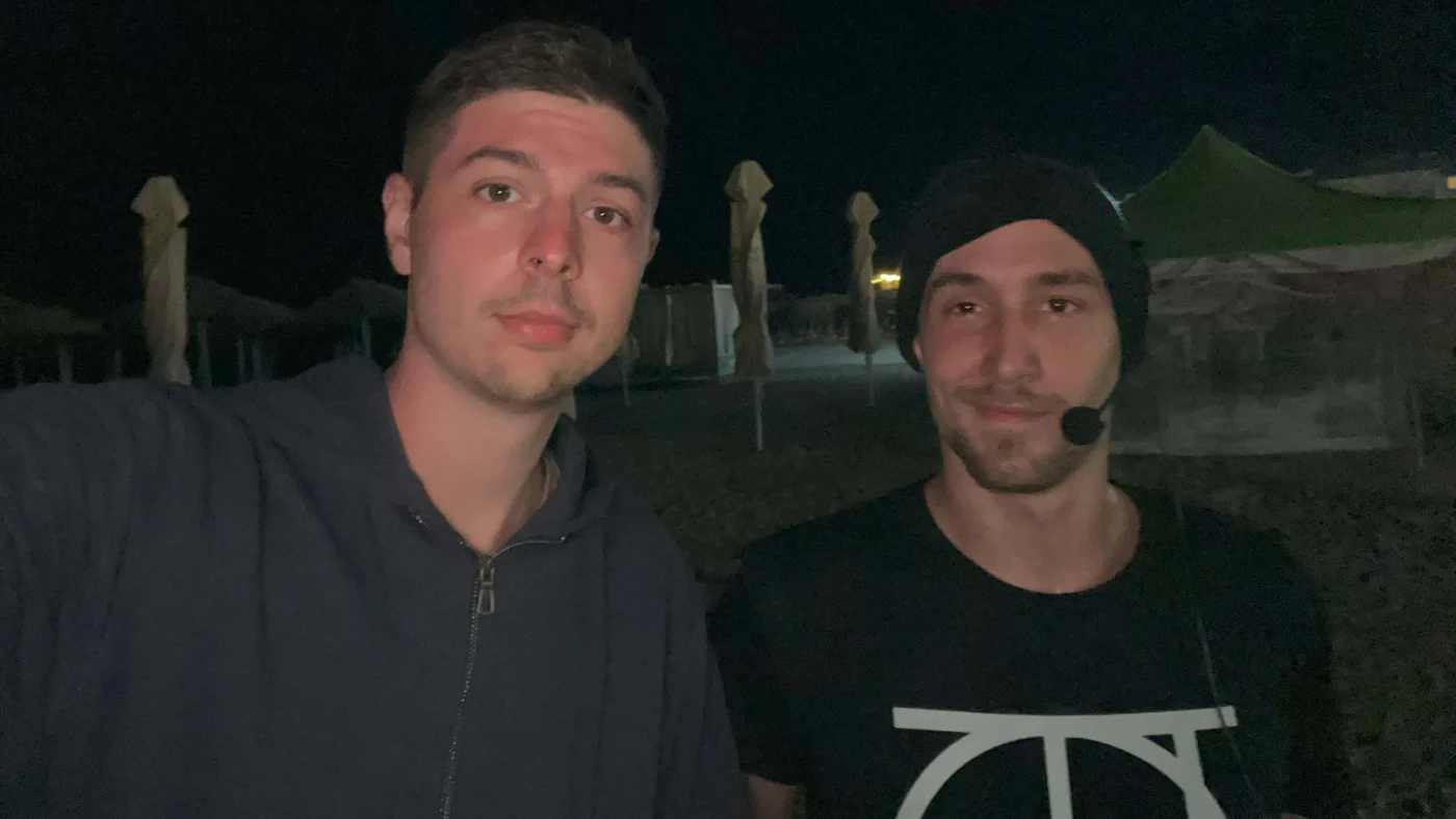 Nighttime selfie on the beach at Vama Veche. Two men shoulder to shoulder; left wears a dark hoodie, right wears a beanie and a headset mic. Folded beach umbrellas and tents sit in the background.