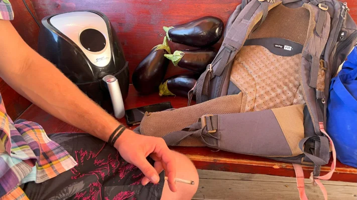 Table scene with a backpack, an air fryer, several eggplants, and a hand holding a cigarette.