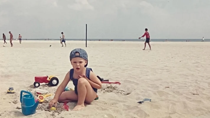 A young boy wearing a backward cap sits on the sandy beach at Vama Veche around 1995–1996, playing with sand toys like a watering can, shovel, and toy truck, while beachgoers walk and play in the background near the sea.