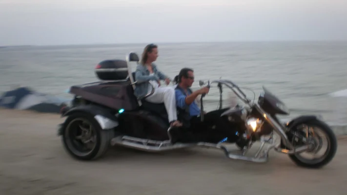 A custom-built trike motorcycle with two riders cruises along the seafront road at dusk.