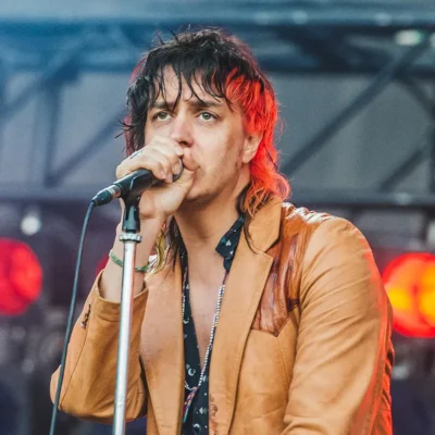 Julian Casablancas performing on stage, holding a microphone, wearing a tan leather jacket.