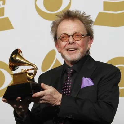 Paul Williams holding a Grammy trophy, wearing glasses, black suit, and purple tie.