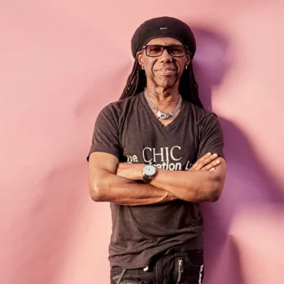 Nile Rodgers smiling in front of a pink background, wearing a black Chic t-shirt and beret.