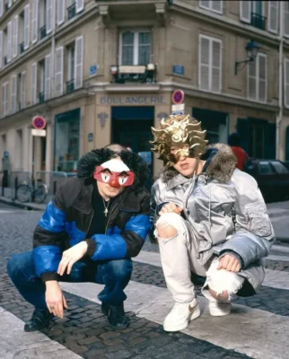 Daft Punk crouching on a Paris street in winter jackets, wearing playful carnival-style masks.