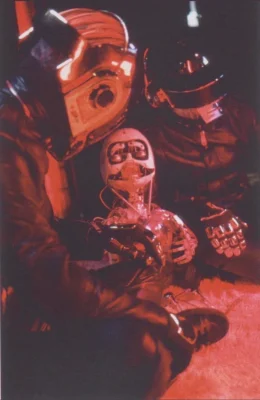 Daft Punk in Human After All helmets and leather jackets, sitting with a robotic mannequin head under red light.